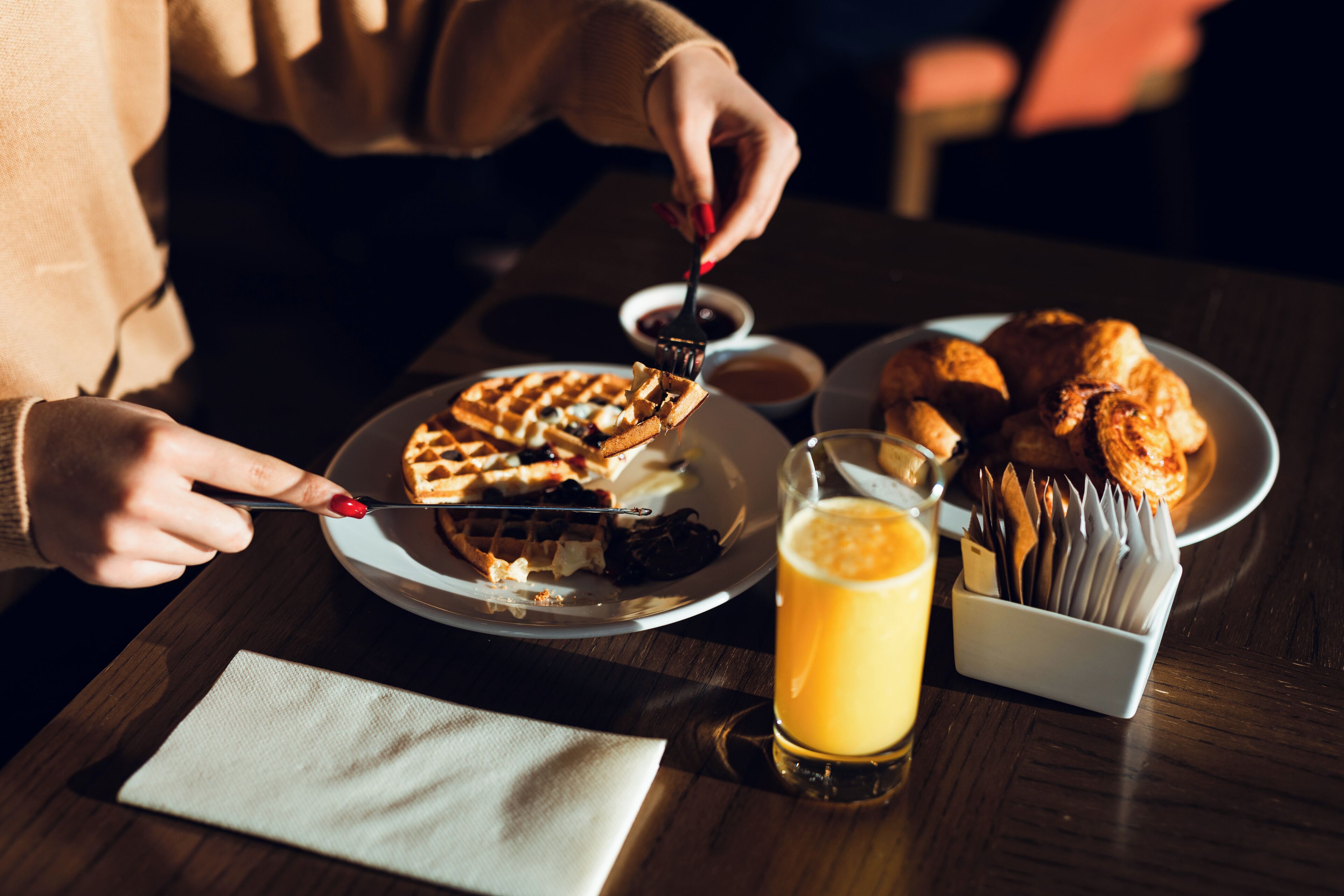 A person eating waffles with syrup, a glass of orange juice, and a plate of croissants on a wooden table.