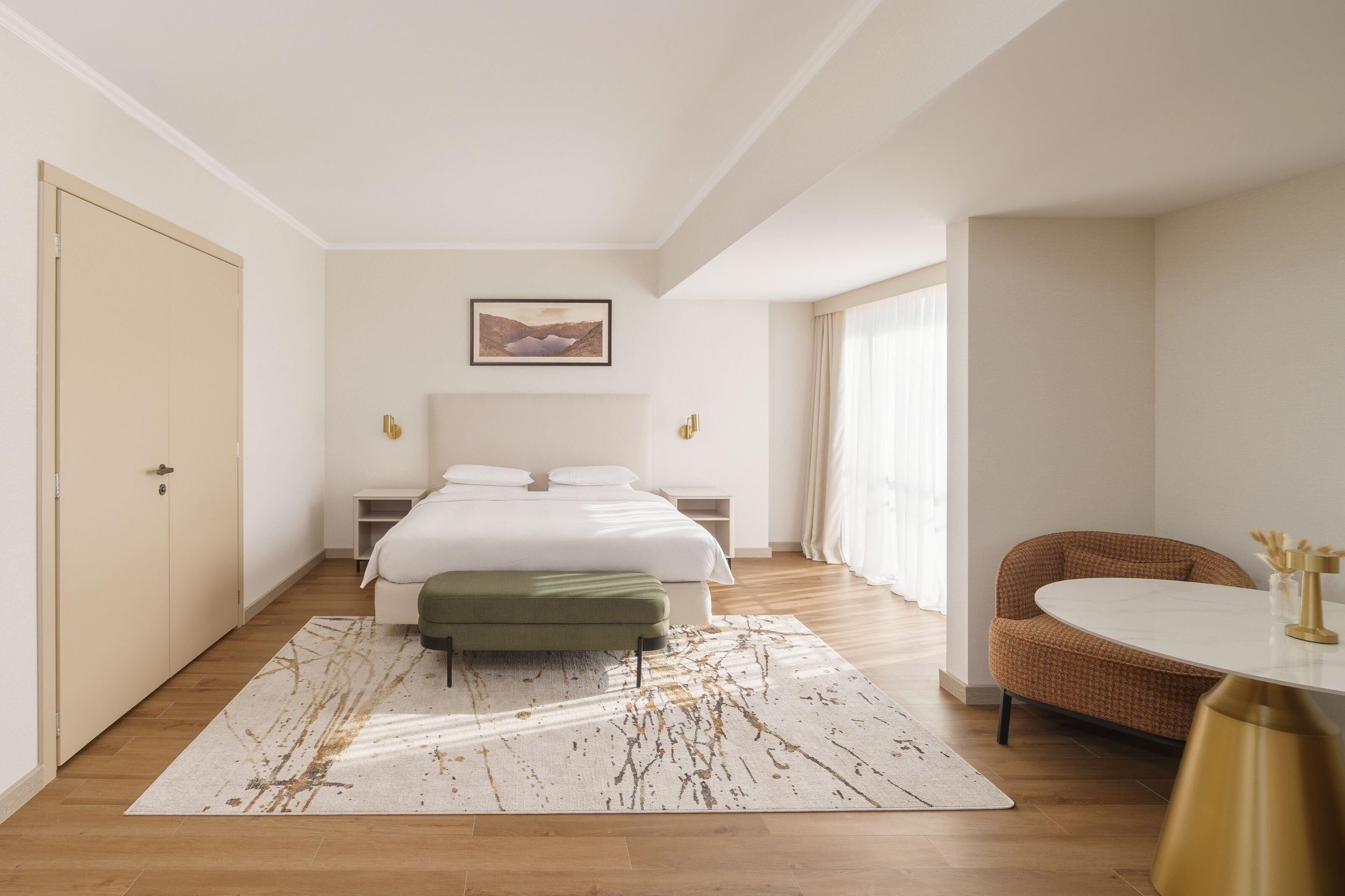 Un dormitorio con cama, mesitas de noche, un banco, una mesa redonda y una silla.