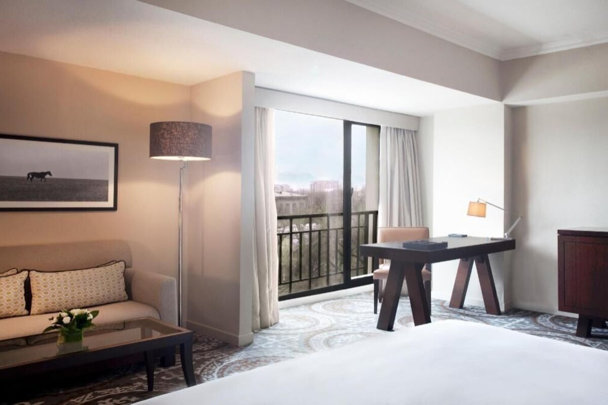 A hotel room with a bed, a sofa, a desk, a lamp, a picture on the wall, and a large window with curtains.