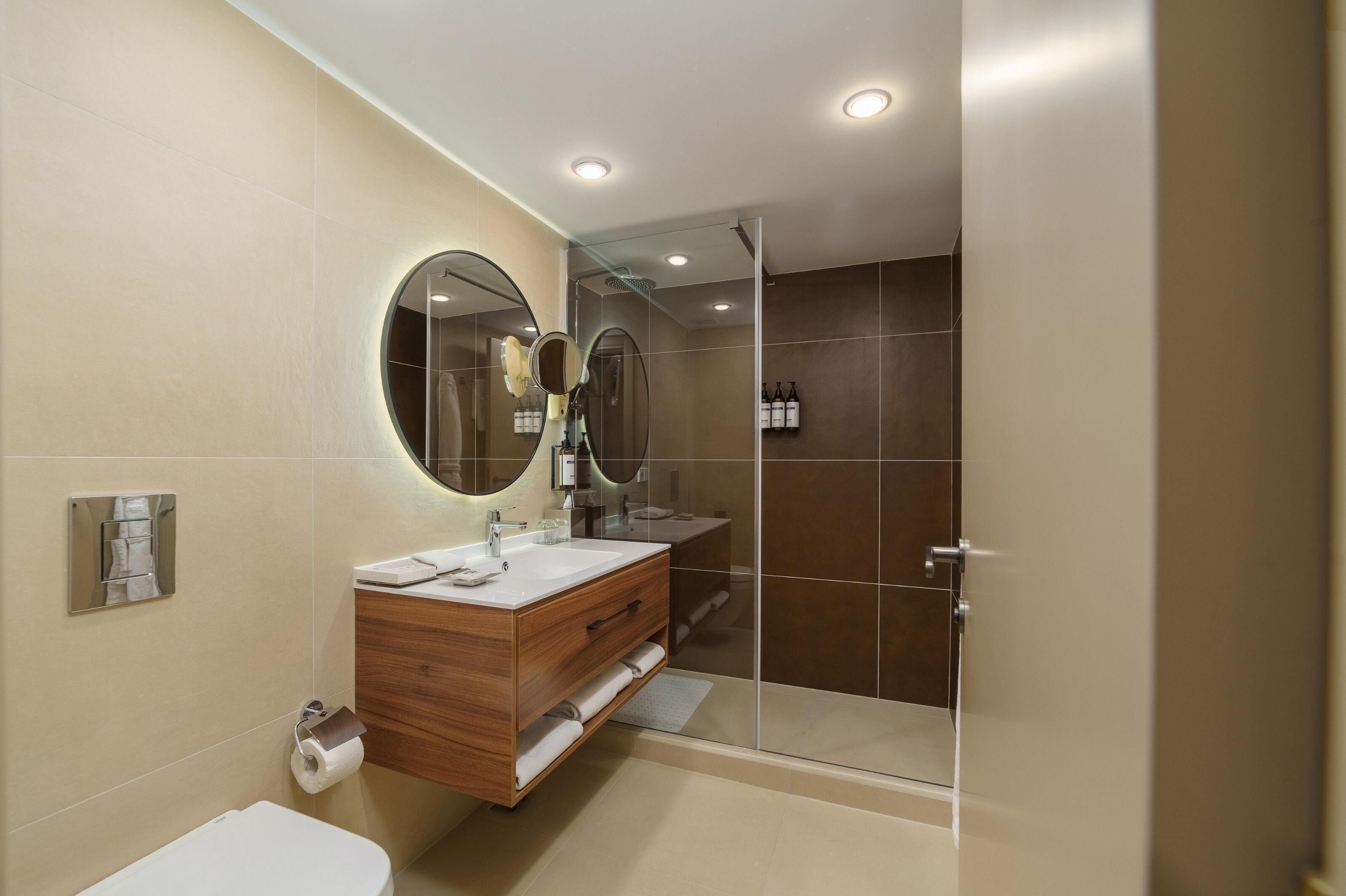 Un baño moderno con tocador de madera, espejo redondo, cabina de ducha de vidrio y retrete.