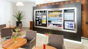 Un moderno vestíbulo de hotel con una estación autoservicio de bebidas, un área de descanso y una planta en maceta.