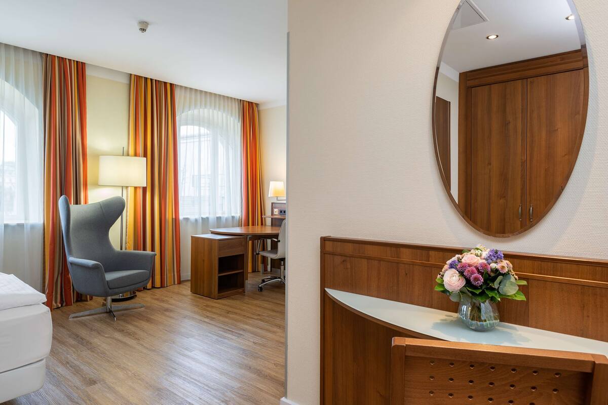 A hotel room with a wooden desk, a grey armchair, a white sofa, and a wooden reception desk with a vase of flowers.