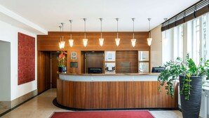Un área de recepción con un mostrador de madera, varias lámparas colgantes, una alfombra roja y una planta grande en maceta.