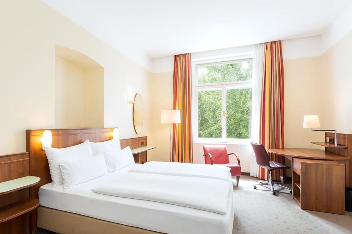 A hotel room with a double bed, a desk, a chair, a lamp, and a window with curtains.