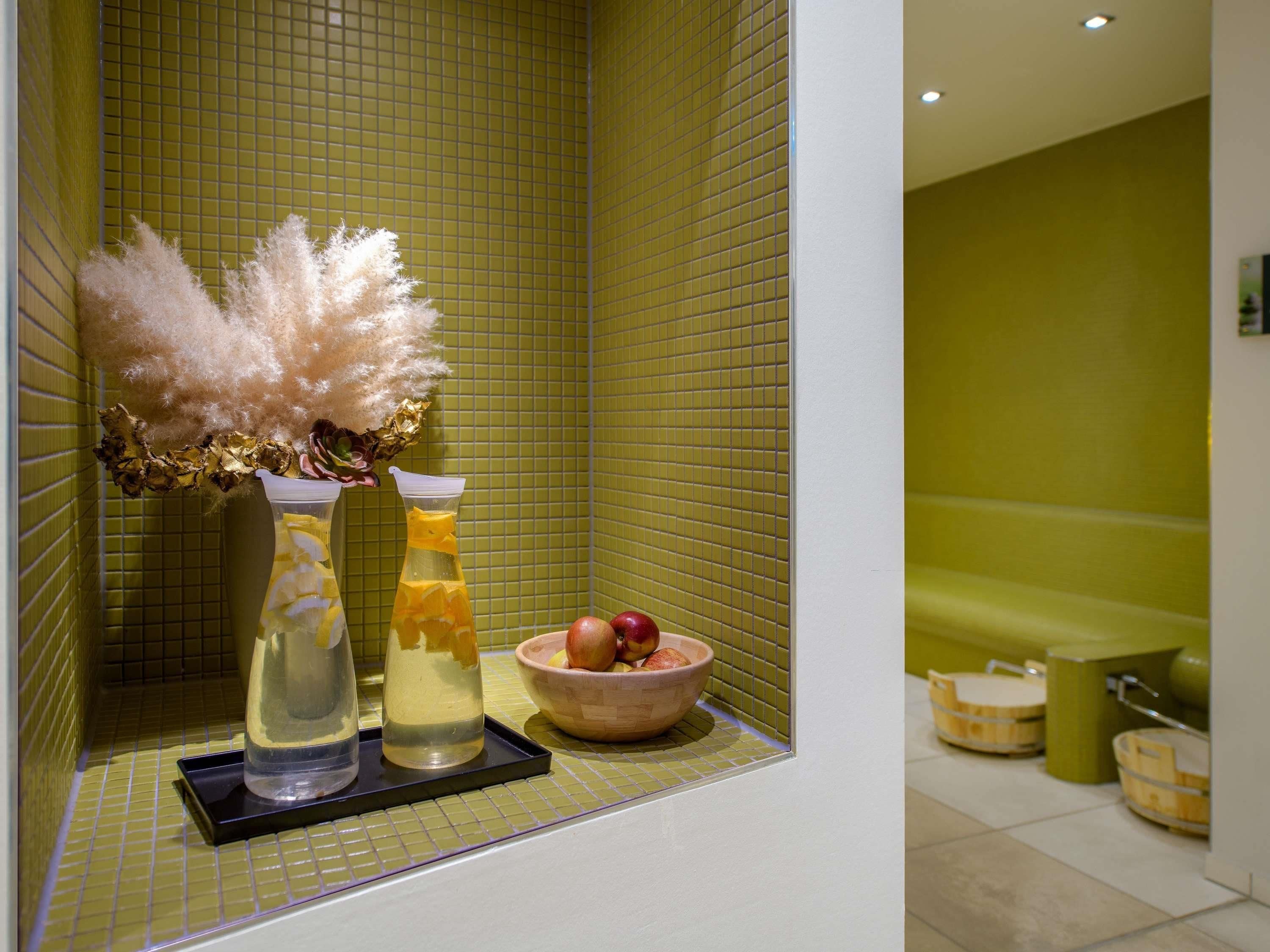 Ein Badezimmer mit einer grünen Fliesenwand, einer Badewanne und einem Regal mit einer Vase getrockneter Pampasgräser, einer Orangensaftflasche und einer Obstschale.