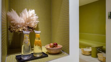 Ein Badezimmer mit einer grünen Fliesenwand, einer Badewanne und einem Regal mit einer Vase getrockneter Pampasgräser, einer Orangensaftflasche und einer Obstschale.