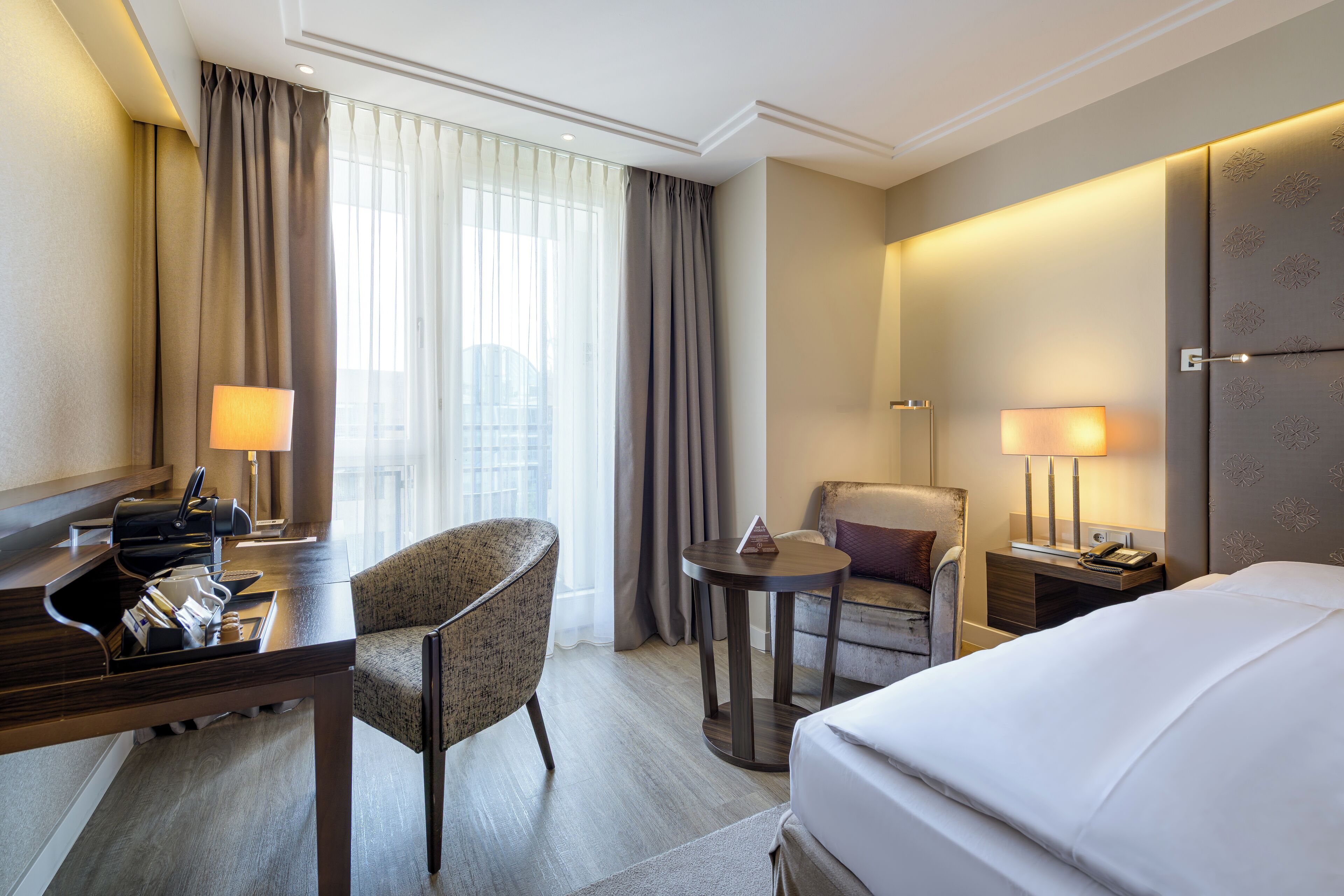 Une chambre d’hôtel comprenant un lit, deux fauteuils, un bureau avec une lampe, une petite table et une table de chevet avec une lampe.