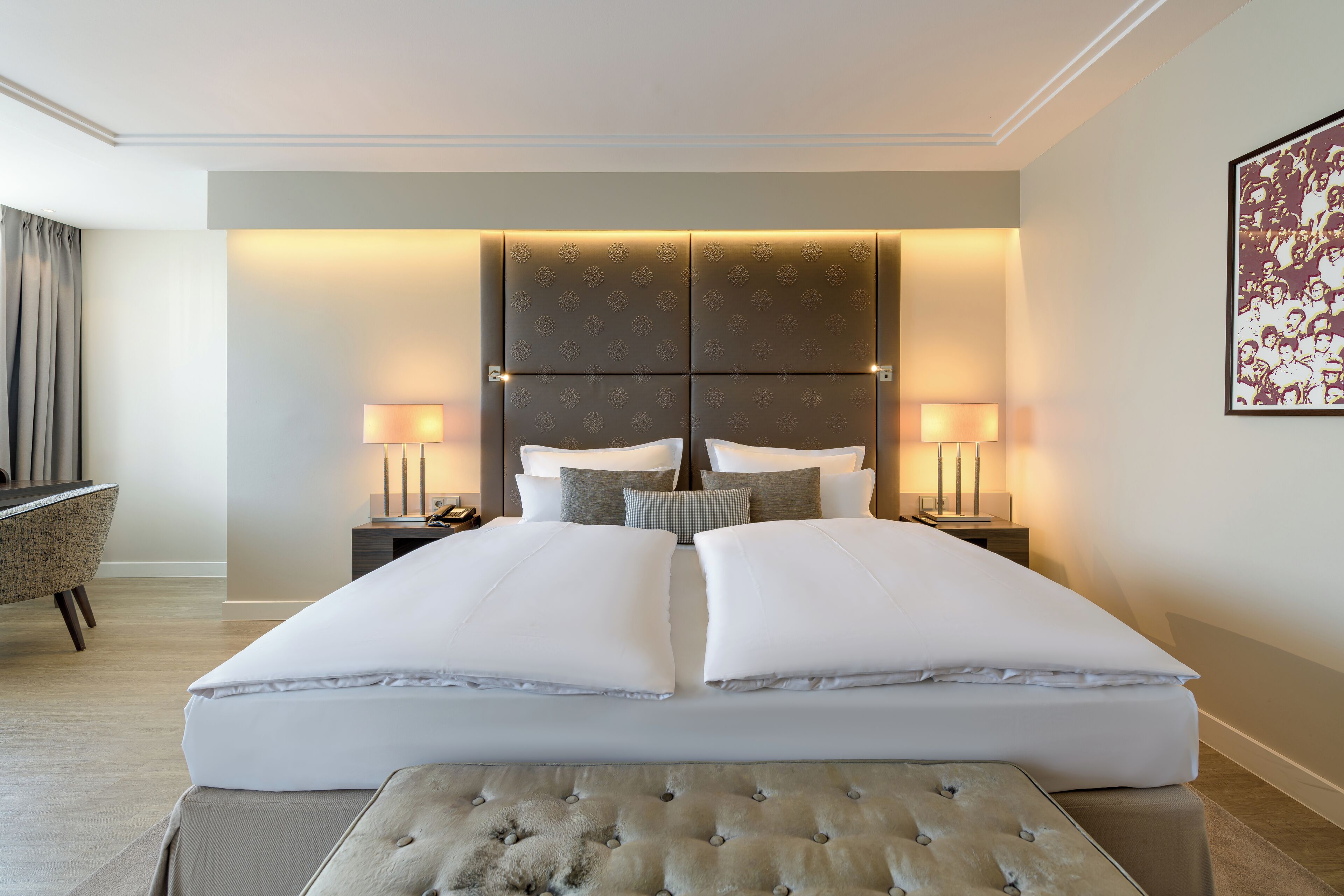 Una cama grande con cabecera capitonada, dos mesitas de noche con lámparas, una silla y una obra de arte enmarcada en la pared.