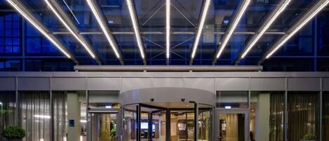 A modern hotel entrance with a revolving door, glass doors, and a paved area with potted plants.
