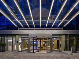 Ingresso moderno di un hotel con porta girevole, vetrate e area pavimentata con piante in vaso.