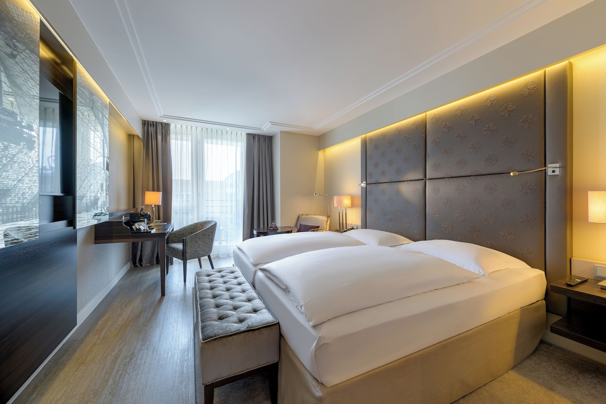 A hotel room with a large bed, a desk, a chair, and a small ottoman.