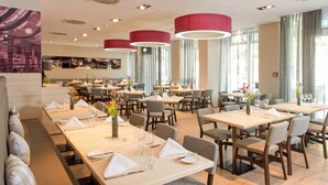 A modern dining area with multiple tables and chairs, large pendant lights, and a view of the cityscape through large windows. - Pullman Munich (Munich)