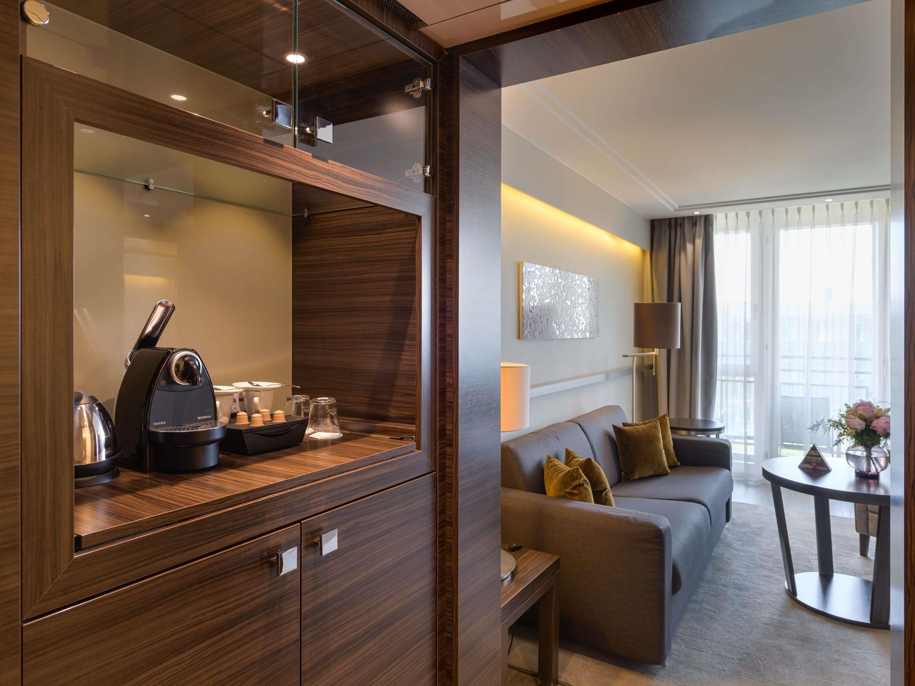A modern hotel room with a wooden vanity, a sofa, a small table, and a large window with curtains.