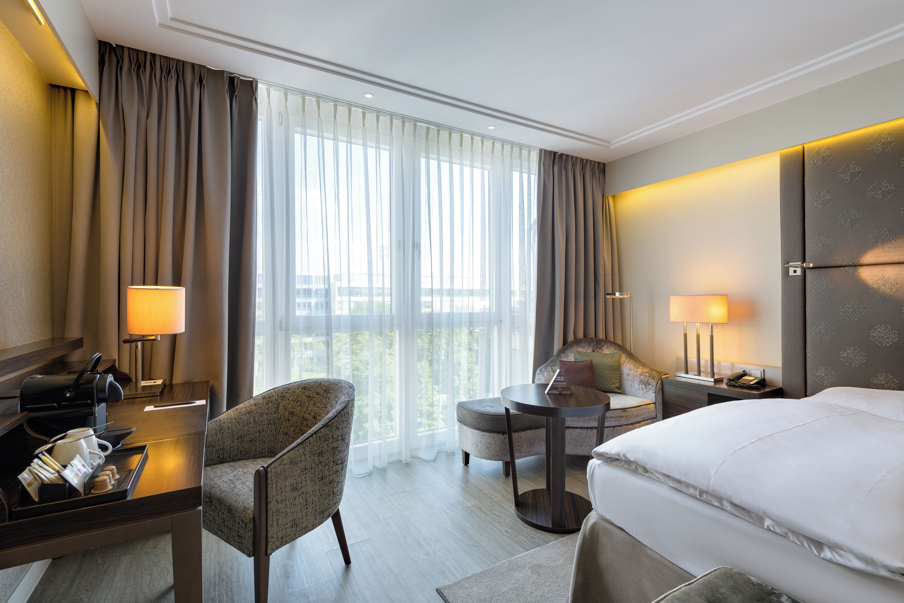 A hotel room with a bed, a desk with a lamp, a chair, a small table, and a view of the outdoors through large windows.