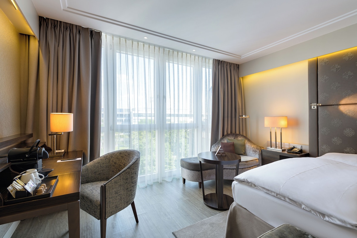 A hotel room with a bed, a desk with a lamp, a chair, a small table, and a view of the outdoors through large windows.