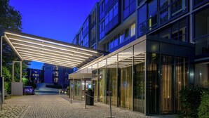 A modern hotel entrance with a covered walkway, glass doors, and a car parked outside. - Pullman Munich (Munich)