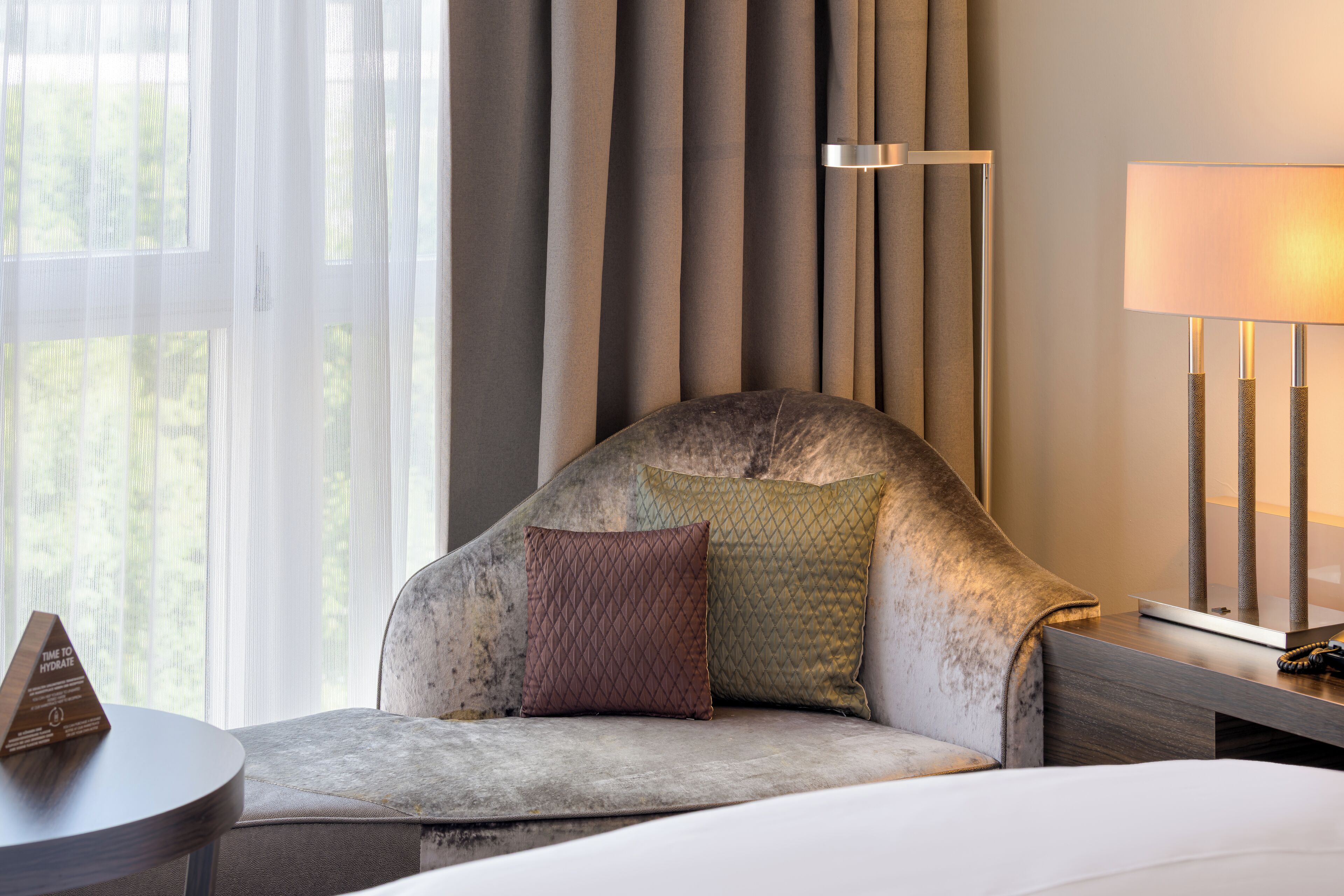 Ein Hotelzimmer mit großem Fenster, einer Couch mit zwei Kissen, einem Nachttisch und einer Lampe.