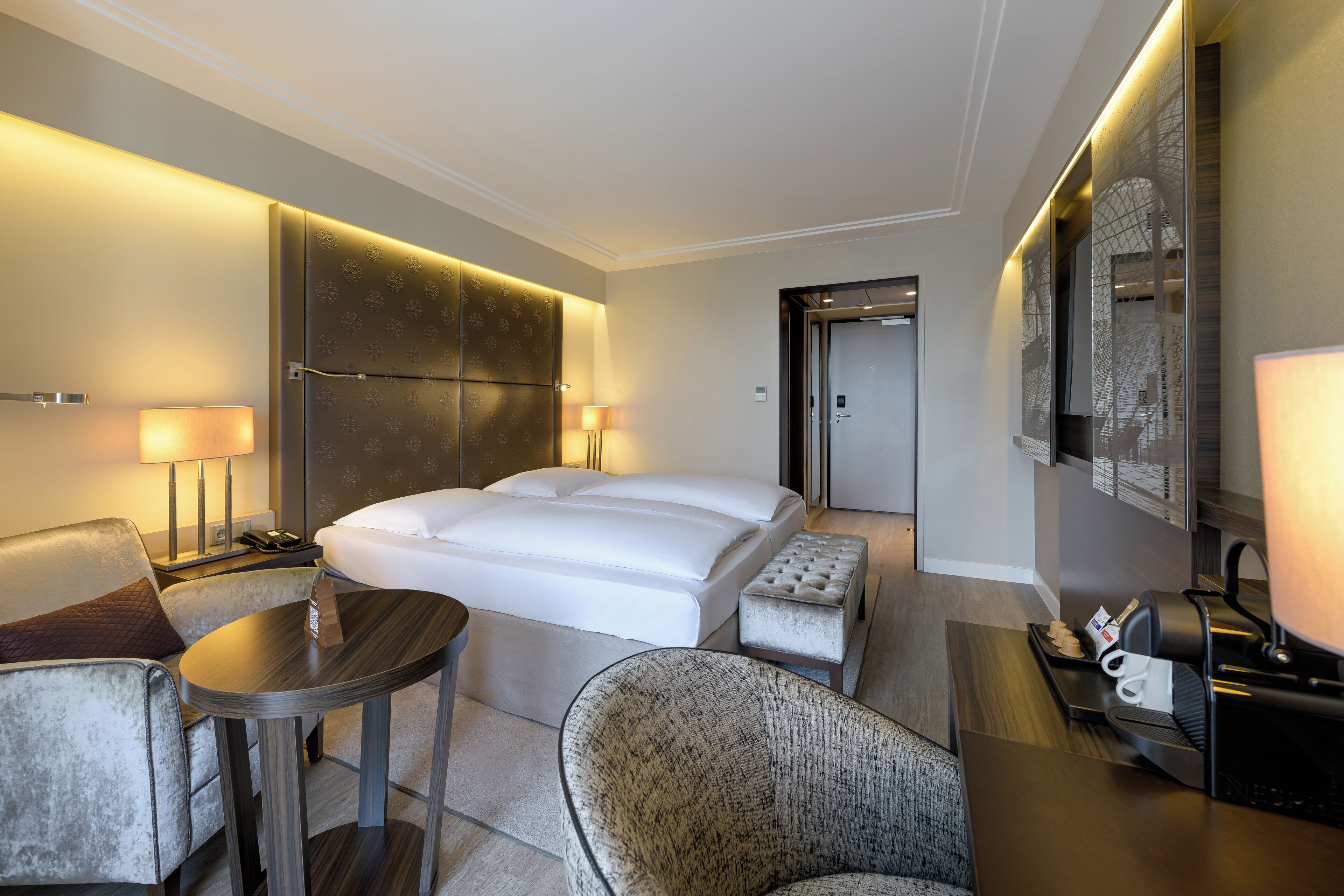 Una habitación de hotel con una cama grande, un área de descanso con una silla y una mesita, un escritorio con una lámpara y un espejo grande en la pared.