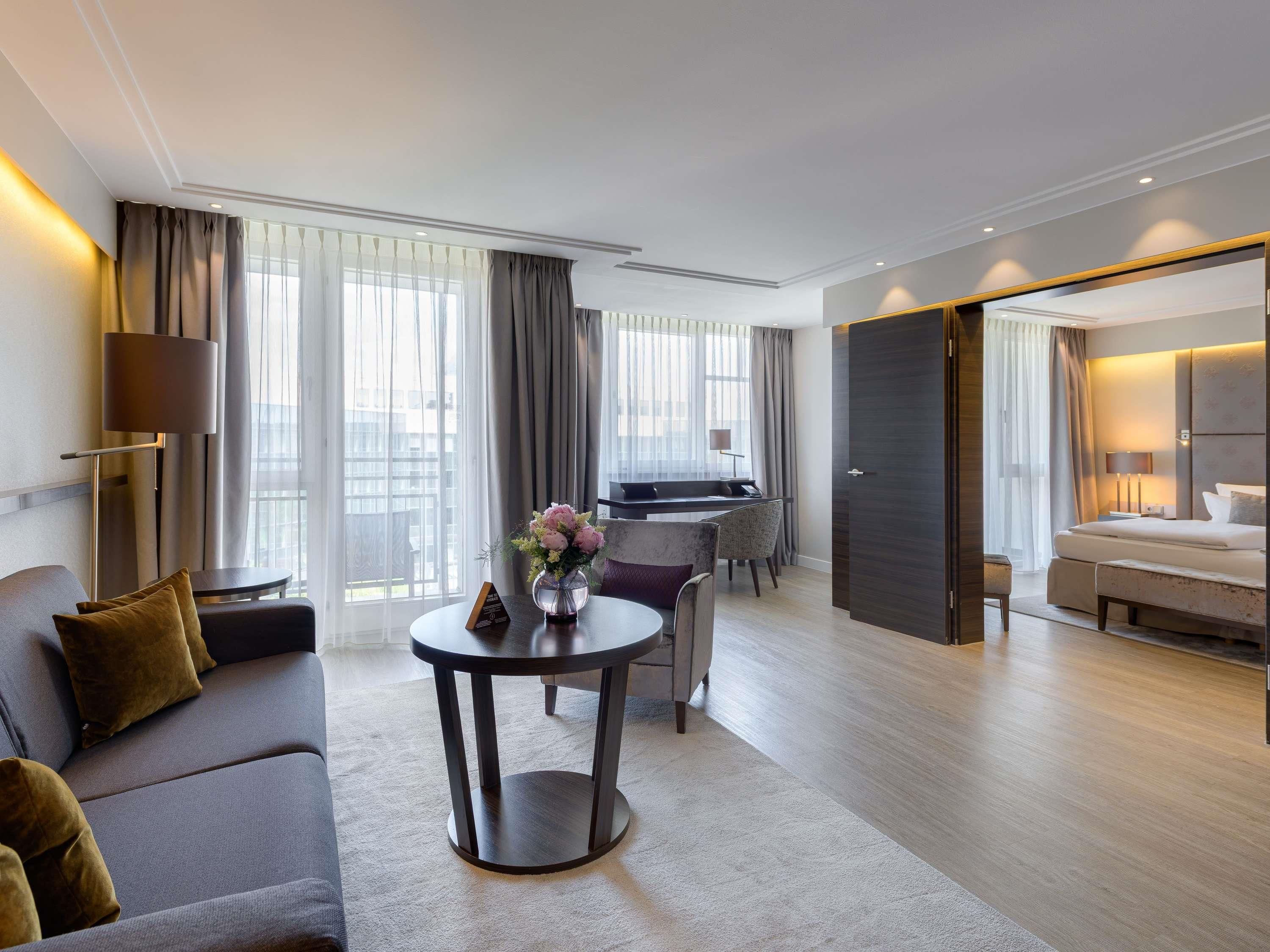 A modern hotel room with a sofa, a round table with a vase, a bed, and a desk area.