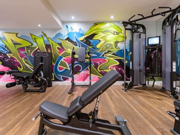 Un gimnasio con equipamiento de ejercicio, una prensa de banca y un mural colorido en la pared.