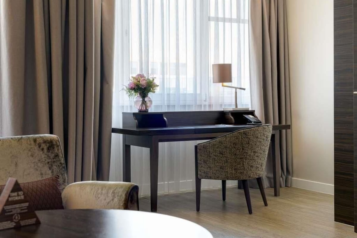 A hotel room with a desk, a chair, a lamp, and a vase with flowers.