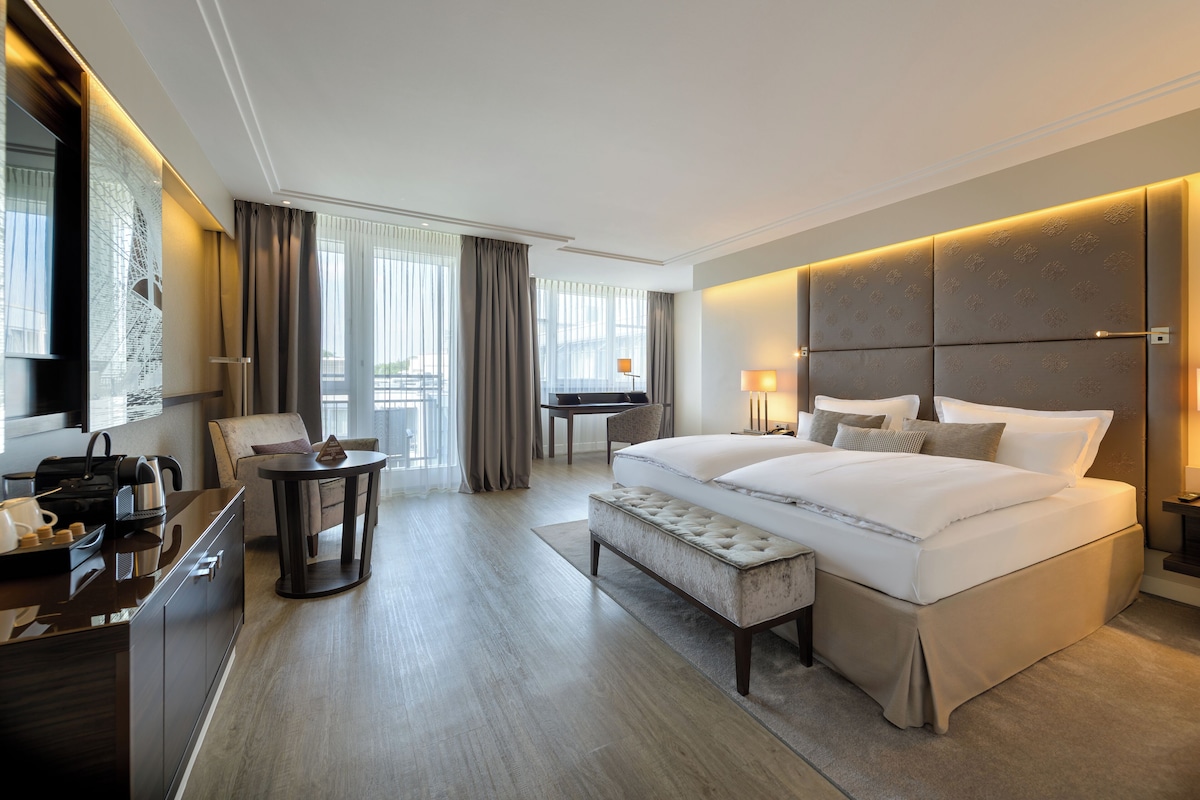 A modern hotel room with a large bed, a desk, a chair, and a view of the outdoors.