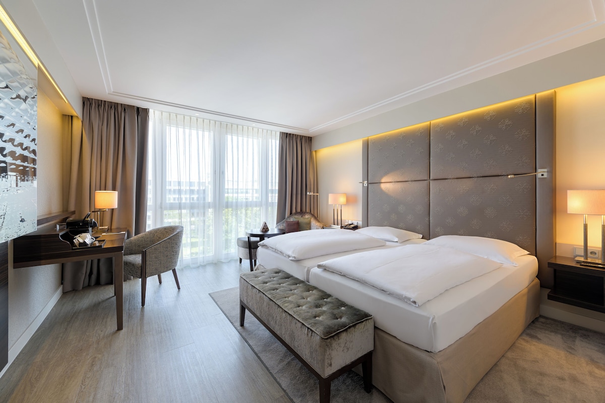 A hotel room with a large bed, a desk, a chair, a bench, and a nightstand with lamps.