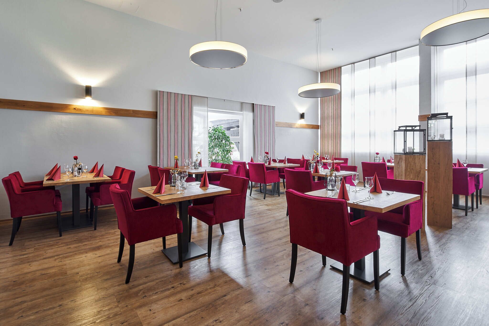 Een restaurant met houten tafels en rode stoelen, grote ramen en moderne hanglampen.