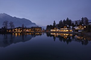 Un hôtel situé au bord d’un lac, composé de plusieurs bâtiments dont les façades se reflètent dans l’eau, et entouré de montagnes.