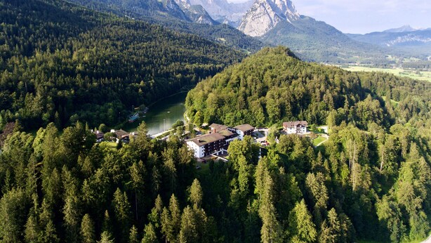 An aerial view of a forested area with a river, a mountain range, and a small cluster of buildings with a lake in the foreground.