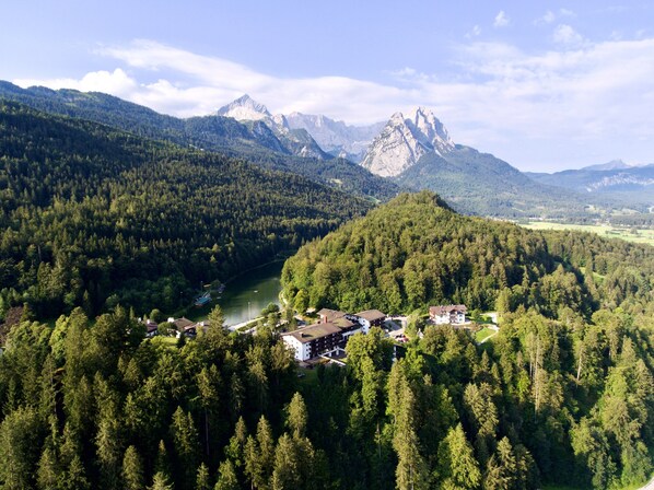 An aerial view of a forested area with a river, a mountain range, and a small cluster of buildings with a lake in the foreground.