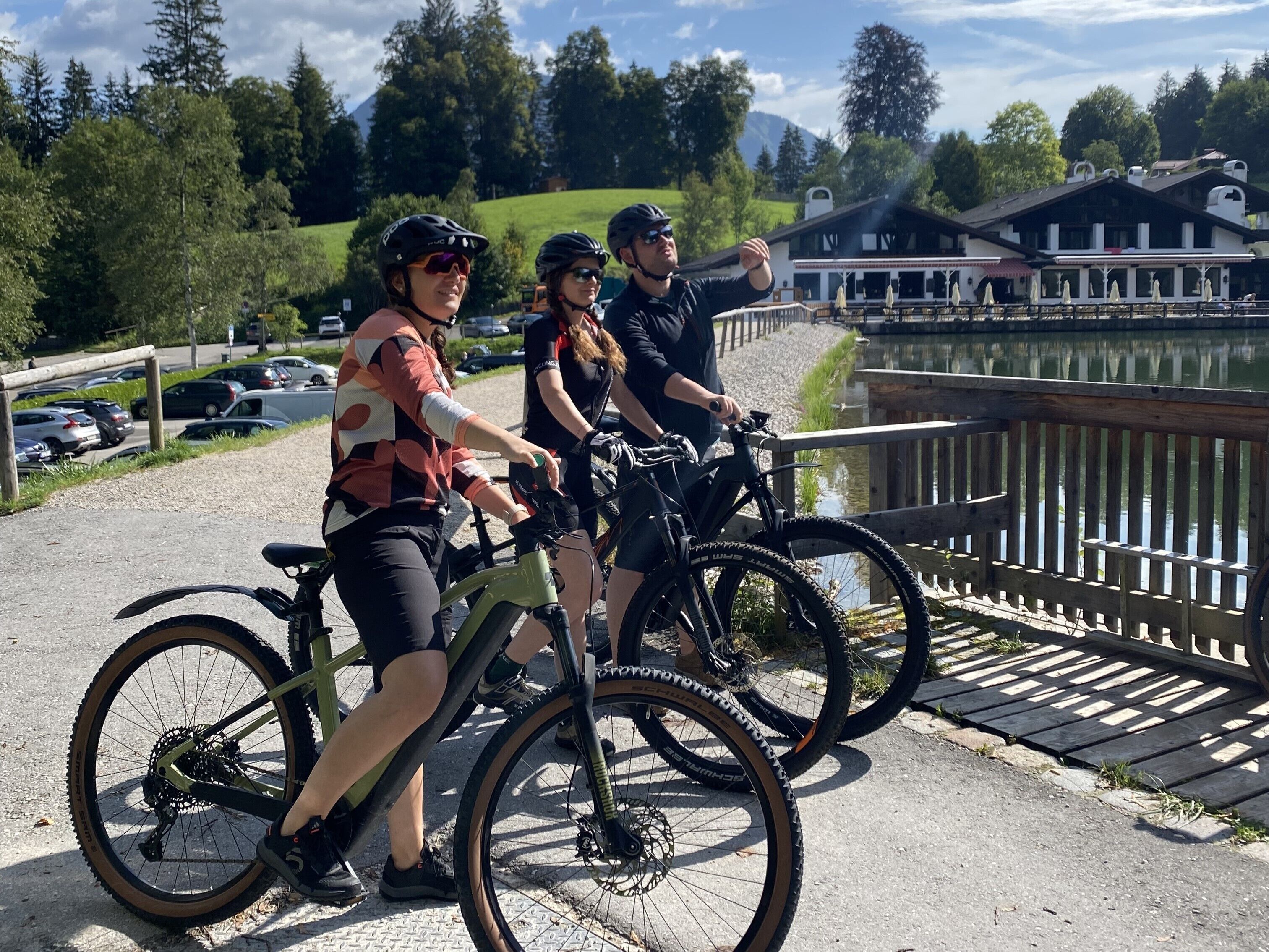 Tre personer på cykler nær en træterrasse ved en sø, med en bygning i baggrunden.