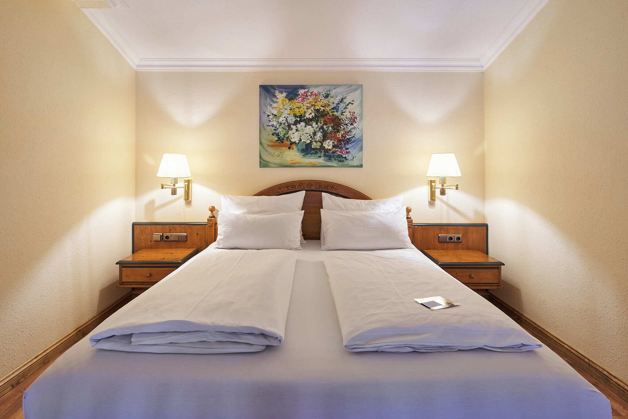 A neatly made double bed with white linens, two bedside tables with lamps, and a floral painting on the wall.