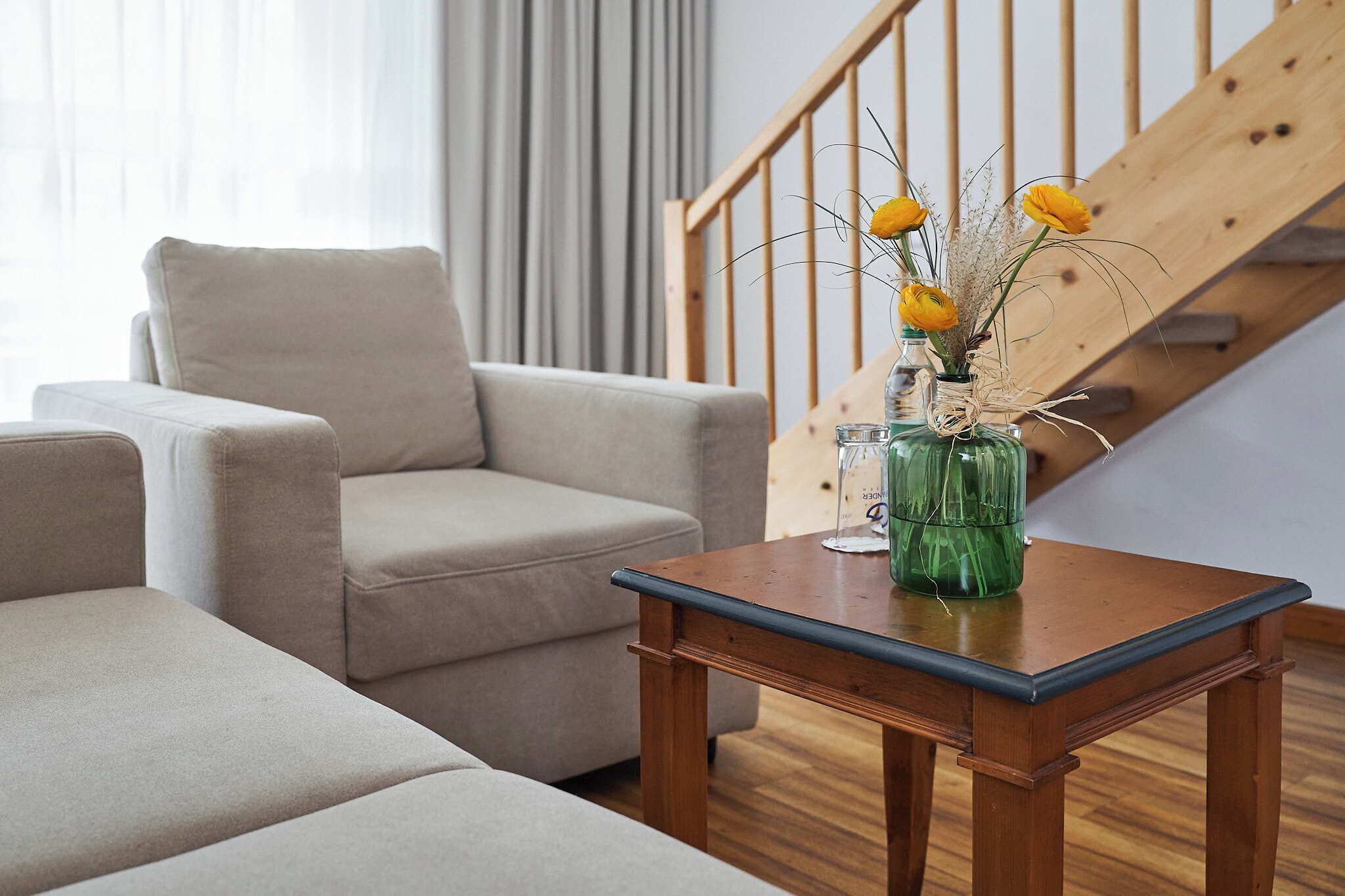 A living room with a beige sofa, a wooden coffee table with a vase of yellow flowers, and a staircase in the background.