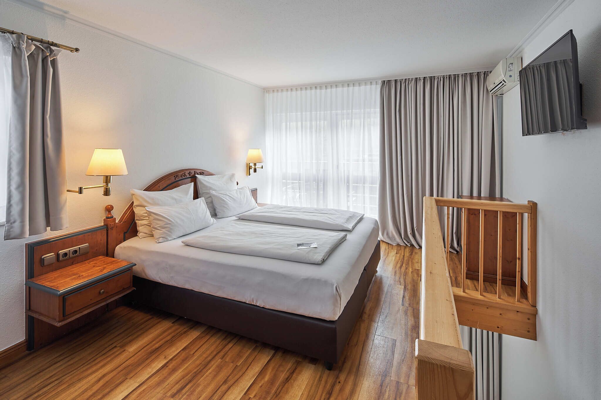 A hotel room with a double bed, bedside lamps, a wooden nightstand, a wooden bench, and a large window with curtains.