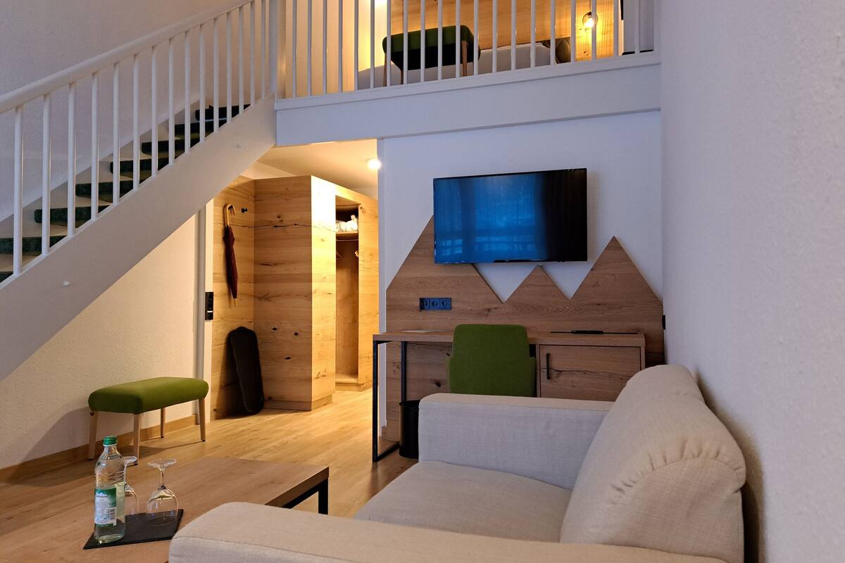 A modern living room with a staircase, a flat-screen TV mounted on a wooden wall, a green chair, and a sofa.