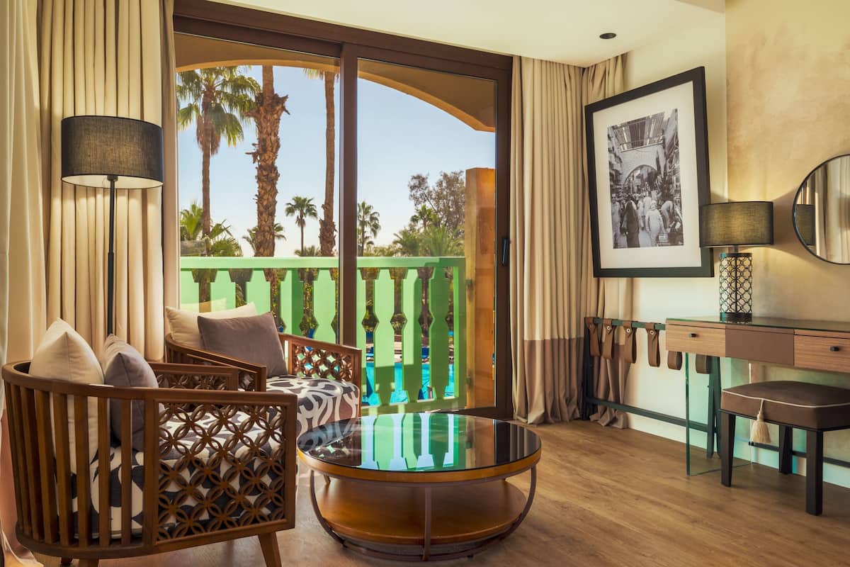 A room with a round glass table, a sofa with patterned cushions, a desk with a lamp, and a framed picture on the wall.