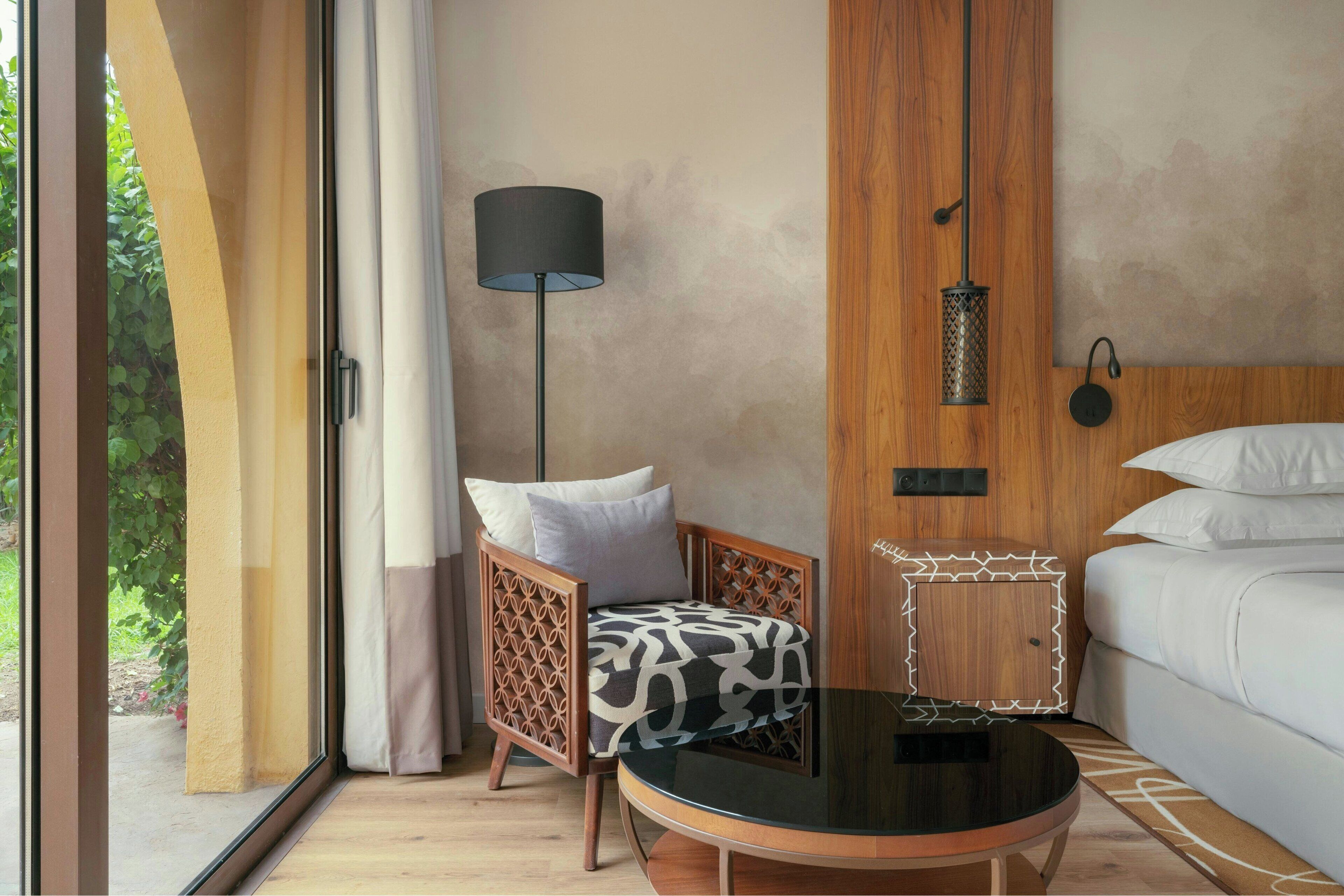 A modern hotel room with a wooden headboard, a round black table, a patterned armchair, and a large window with a view of greenery.