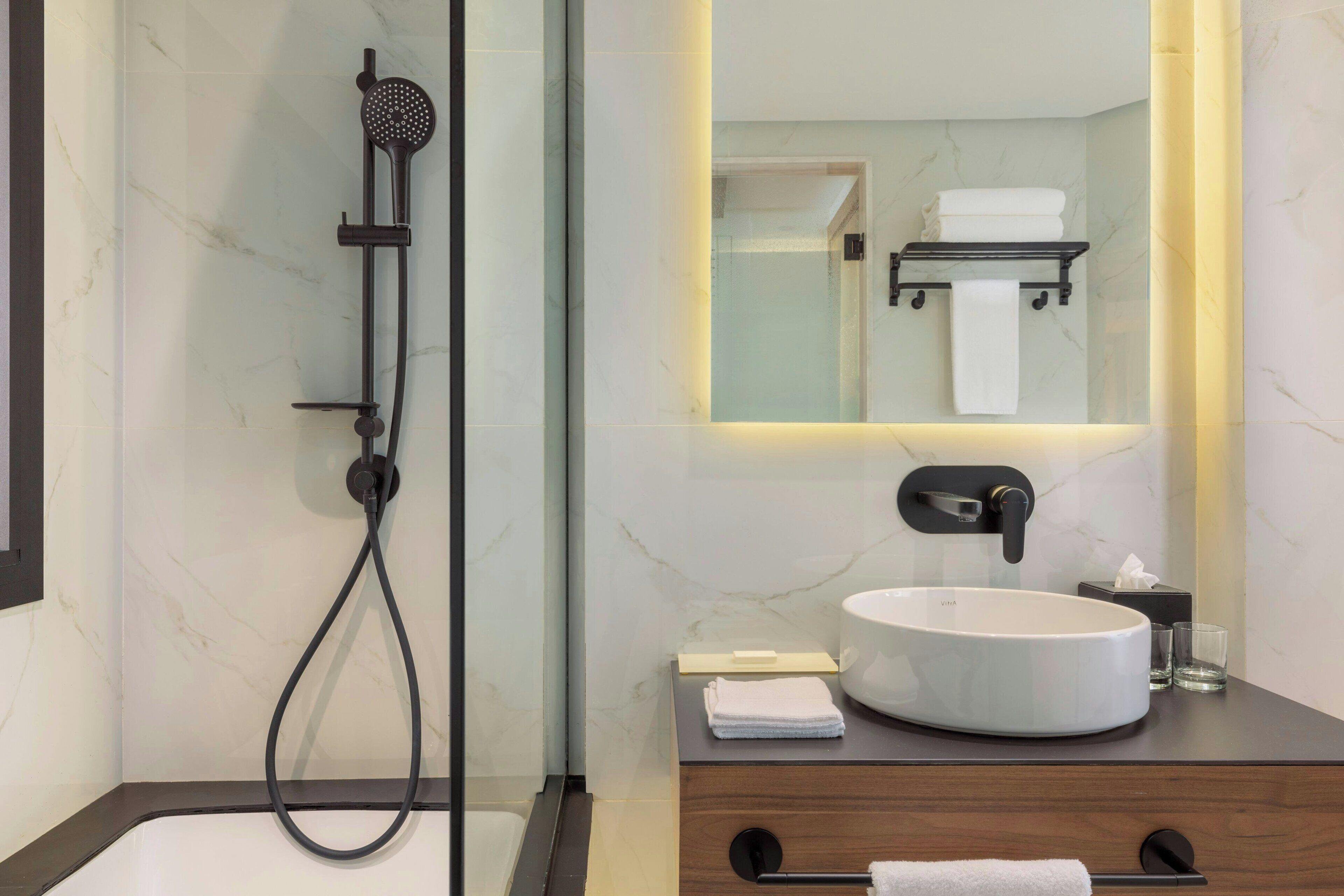 Un baño moderno con ducha envuelta en vidrio, un lavamanos redondo blanco, un tocador de madera oscura y un espejo con toallero.