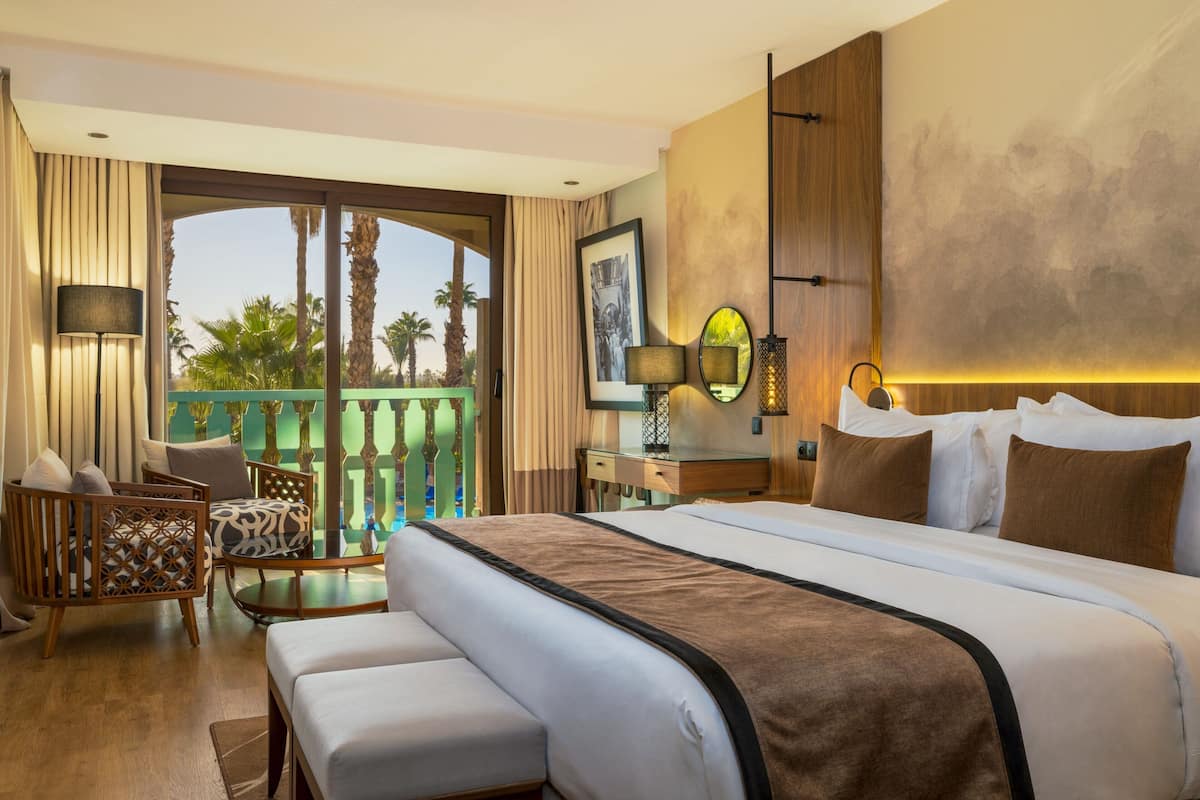 A hotel room with a large bed, a sitting area with a sofa and chairs, a balcony with a view of palm trees, and a wooden accent wall.