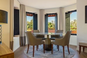 A modern dining area with a round table and six chairs, large windows with curtains, and a view of greenery outside.