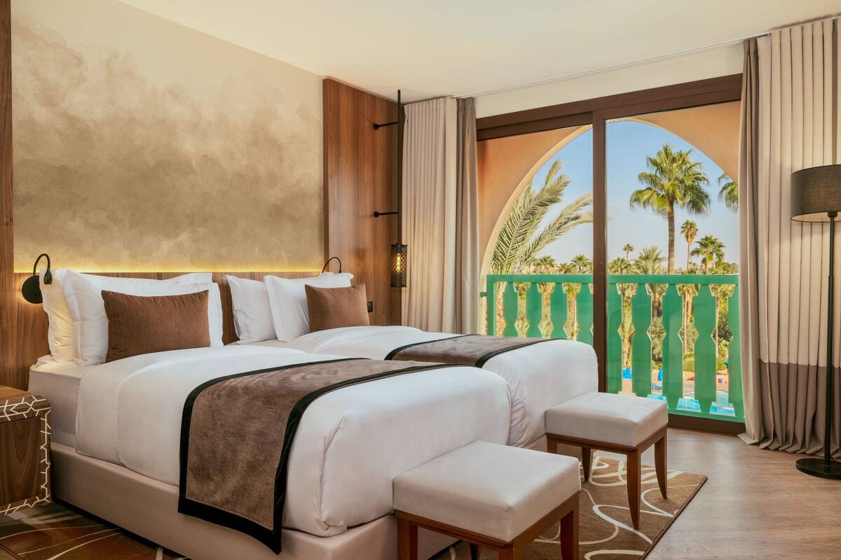 A hotel room with a large bed, a bench, a nightstand, and a view of palm trees through a balcony.