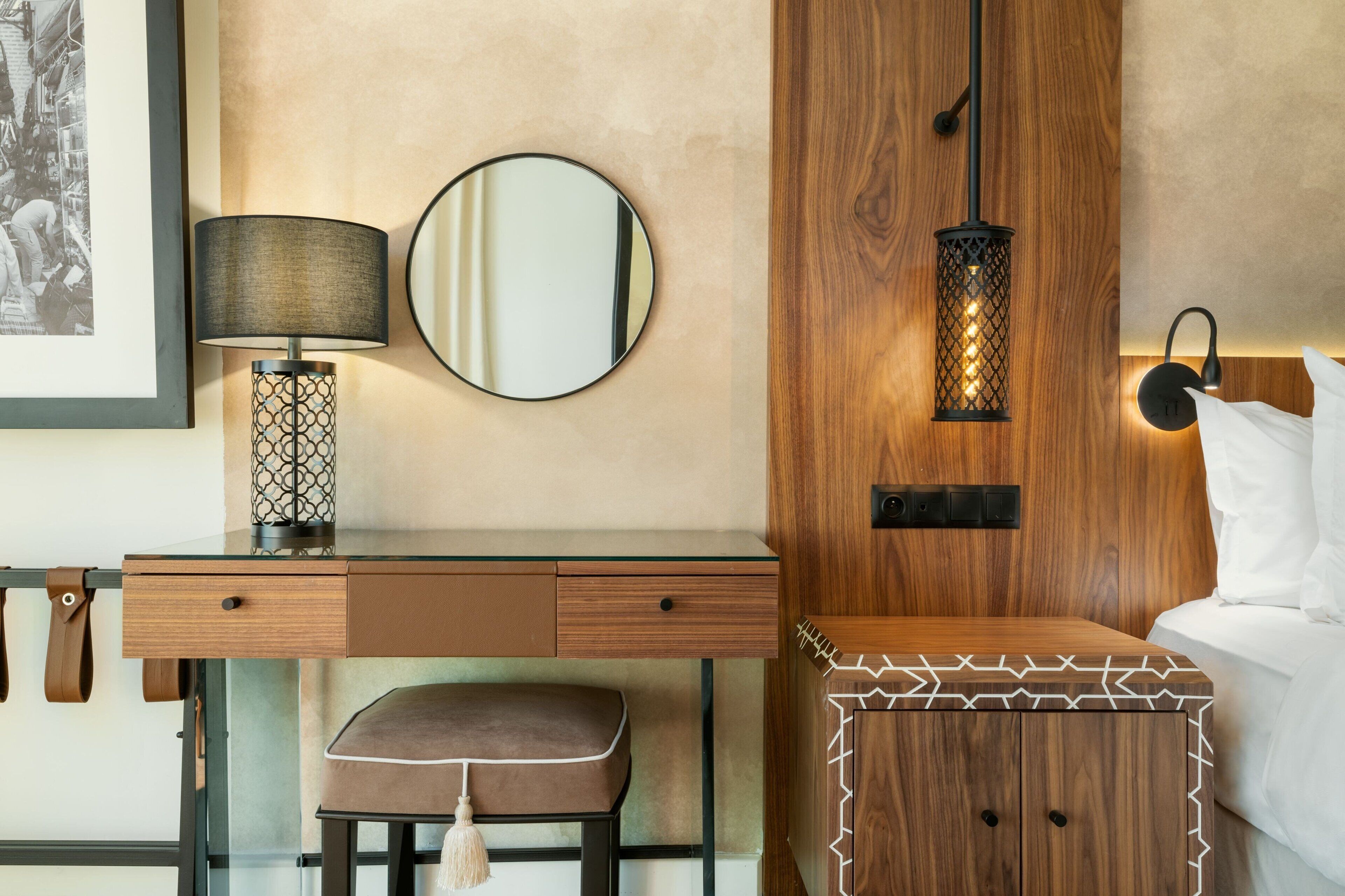 A modern hotel room with a wooden desk, a round mirror, a wall-mounted lamp, and a bedside table with a geometric pattern.