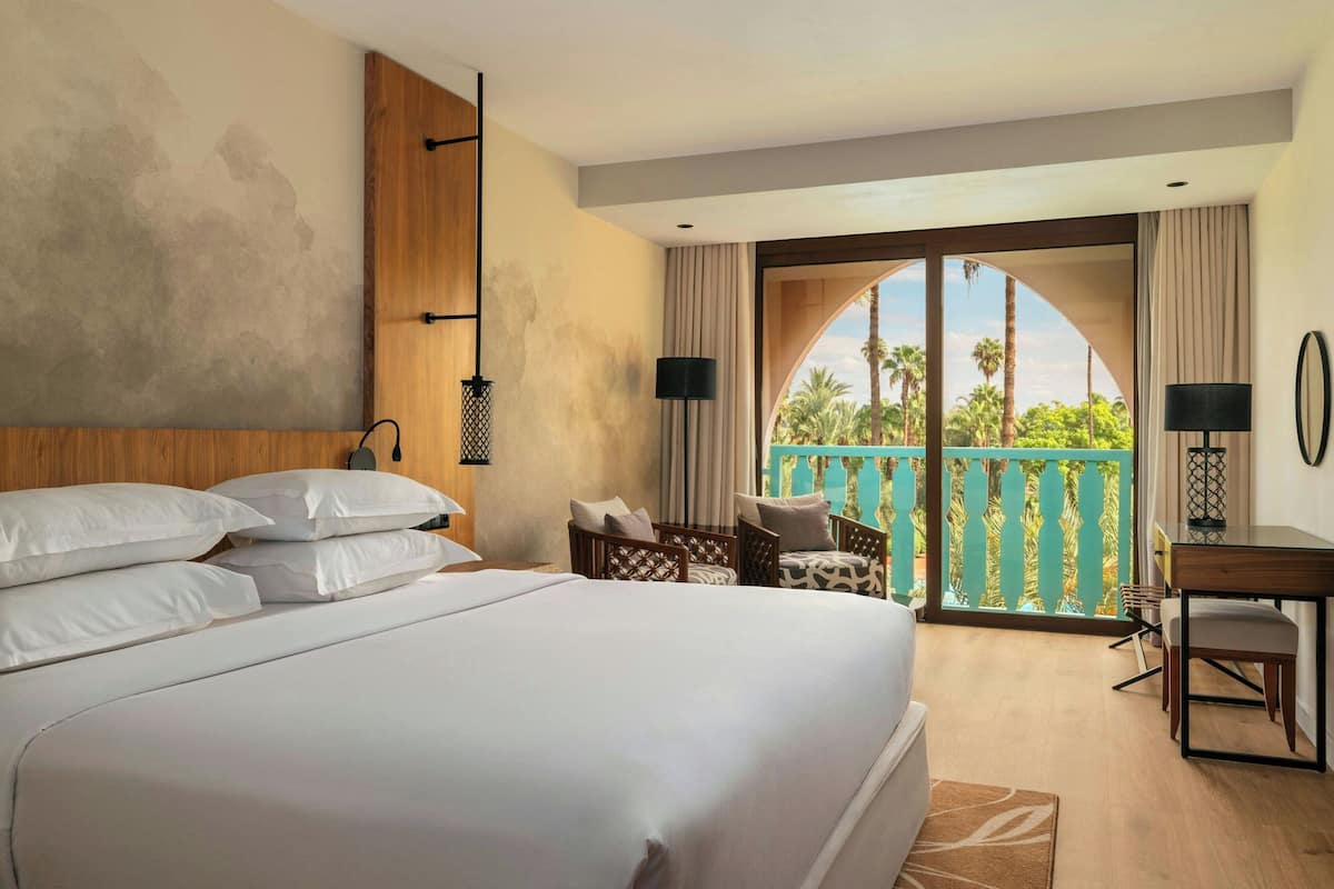A hotel room with a large bed, a desk, a chair, and a balcony with a view of palm trees.