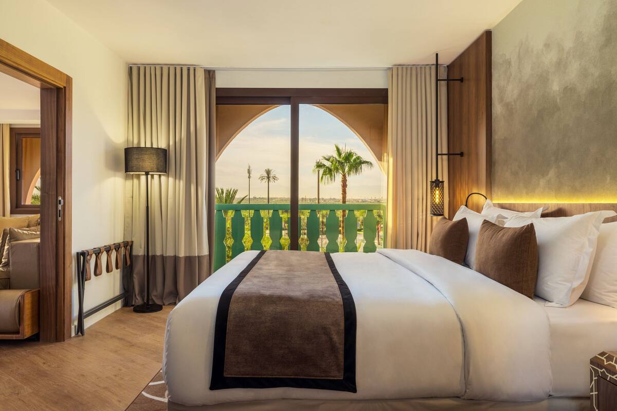 A hotel room with a large bed, a view of palm trees and a clear sky, a balcony with a railing, and a wooden floor.