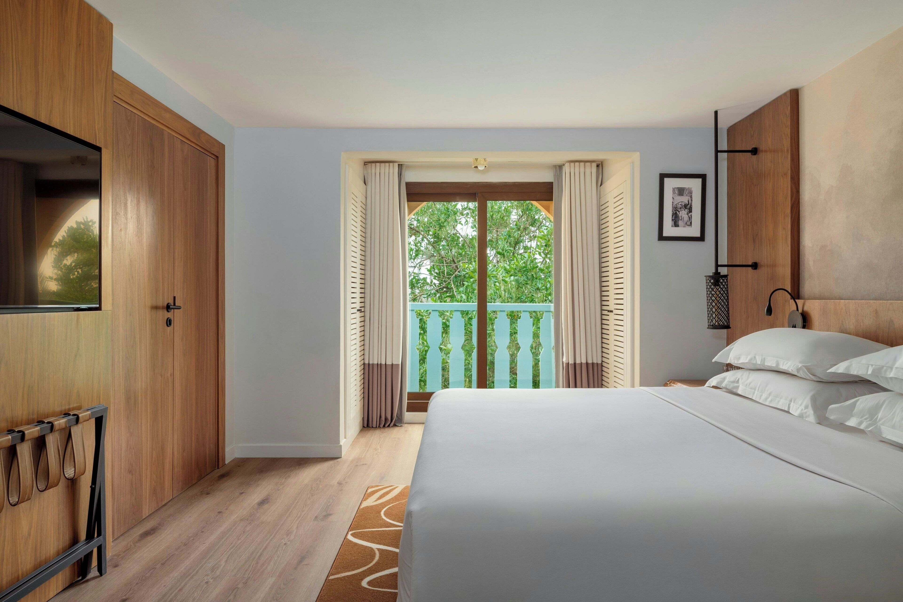 A bedroom with a large bed, wooden wardrobe, and a balcony with a view of greenery.