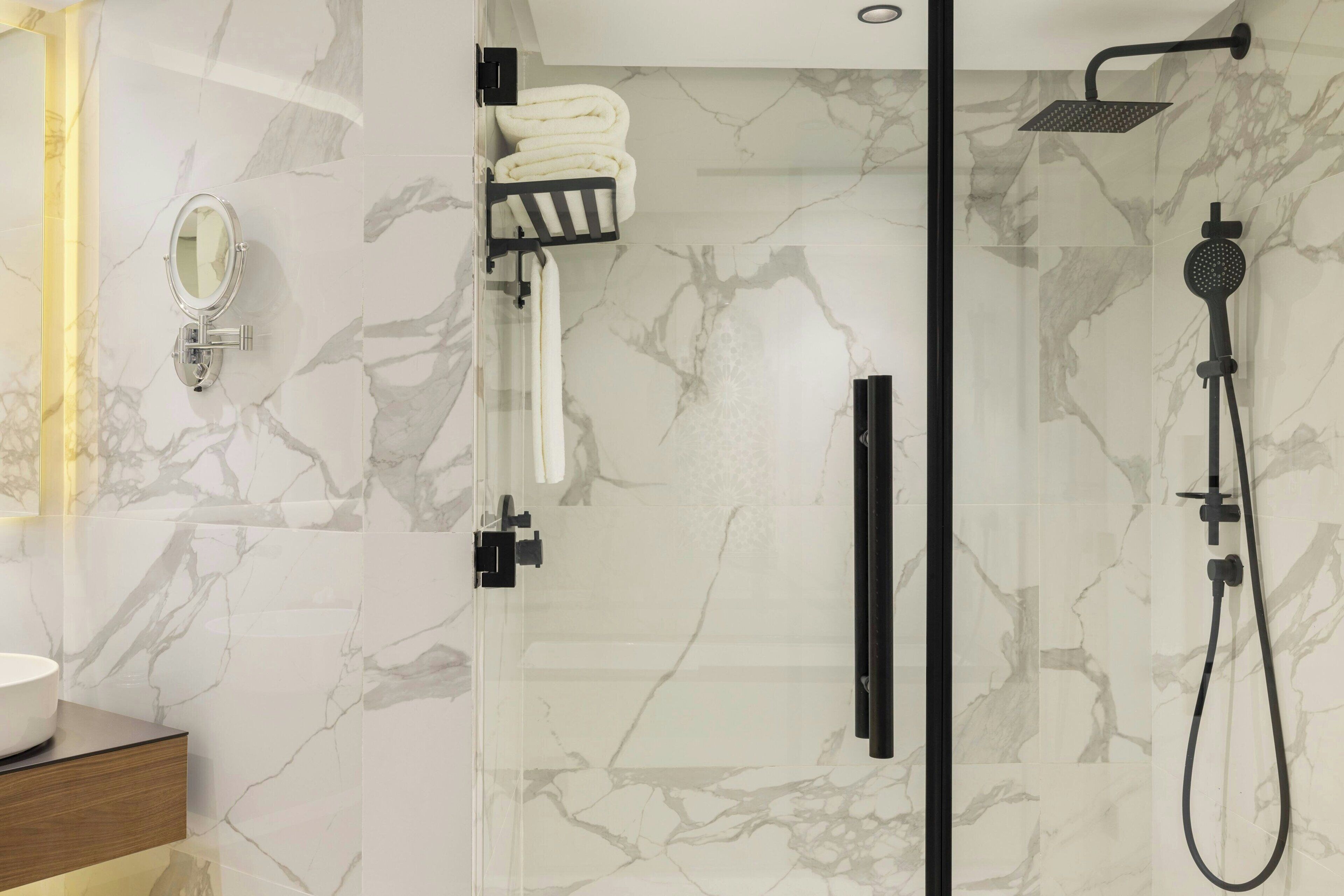 A modern bathroom with a glass shower enclosure, a marble-tiled wall, a white sink, and a towel rack.