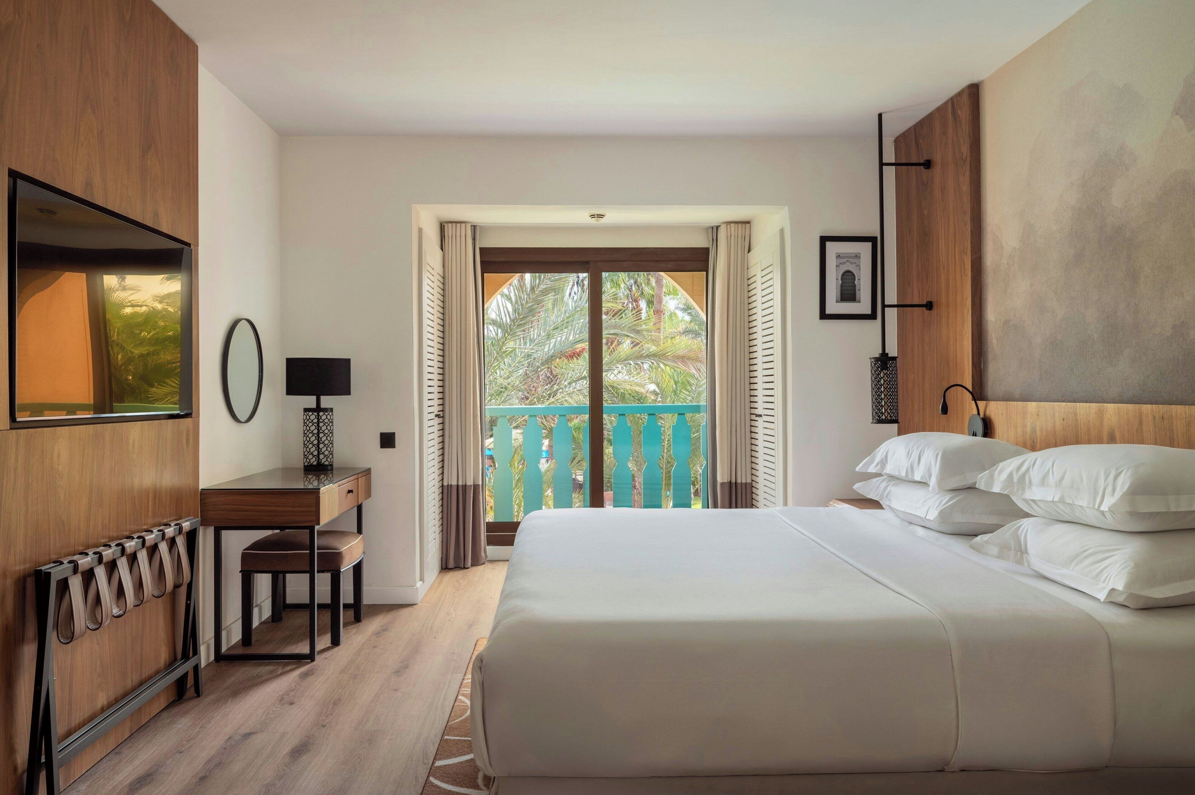 Habitación de hotel con cama, escritorio, silla, espejo y vista a un balcón con barandilla de madera.