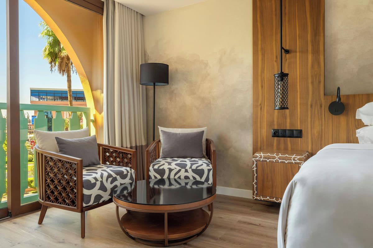 A hotel room with a large window, a round table with two chairs, a bed, and a wooden headboard.