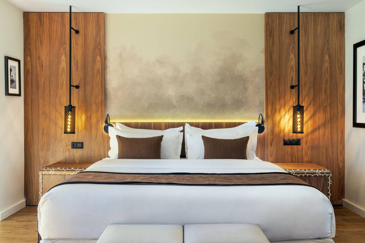 A modern hotel room with a large bed, wooden headboard, and two wall-mounted lamps.